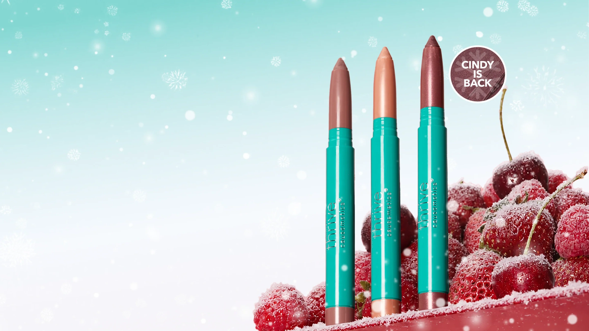 Three turquoise Thrive Causemetics lip sticks standing beside frosted red cherries
